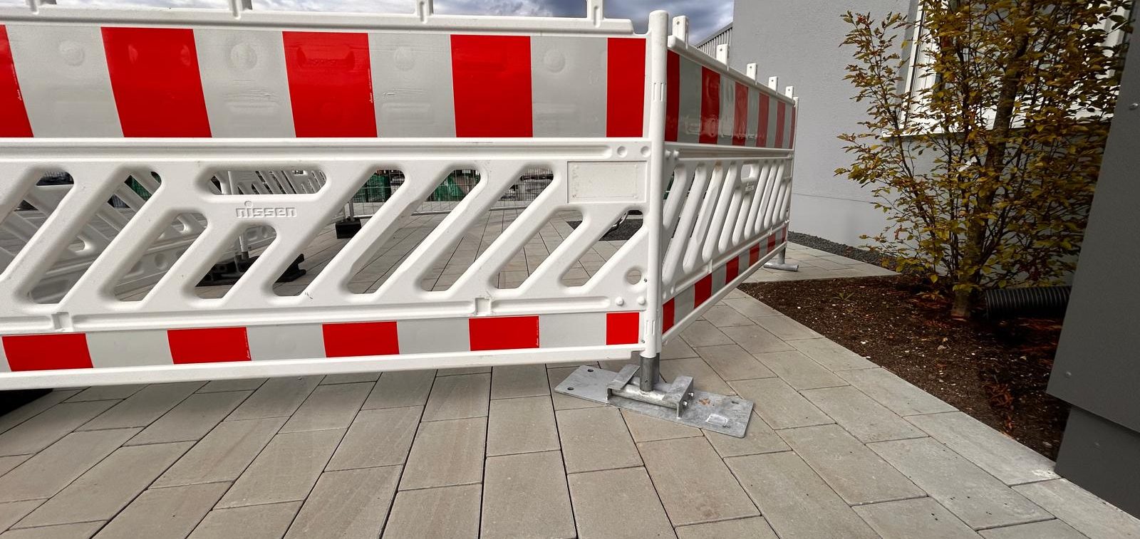 Bauzaun mit roten und weißen Streifen steht auf einem Gehweg neben einer Pflanze.