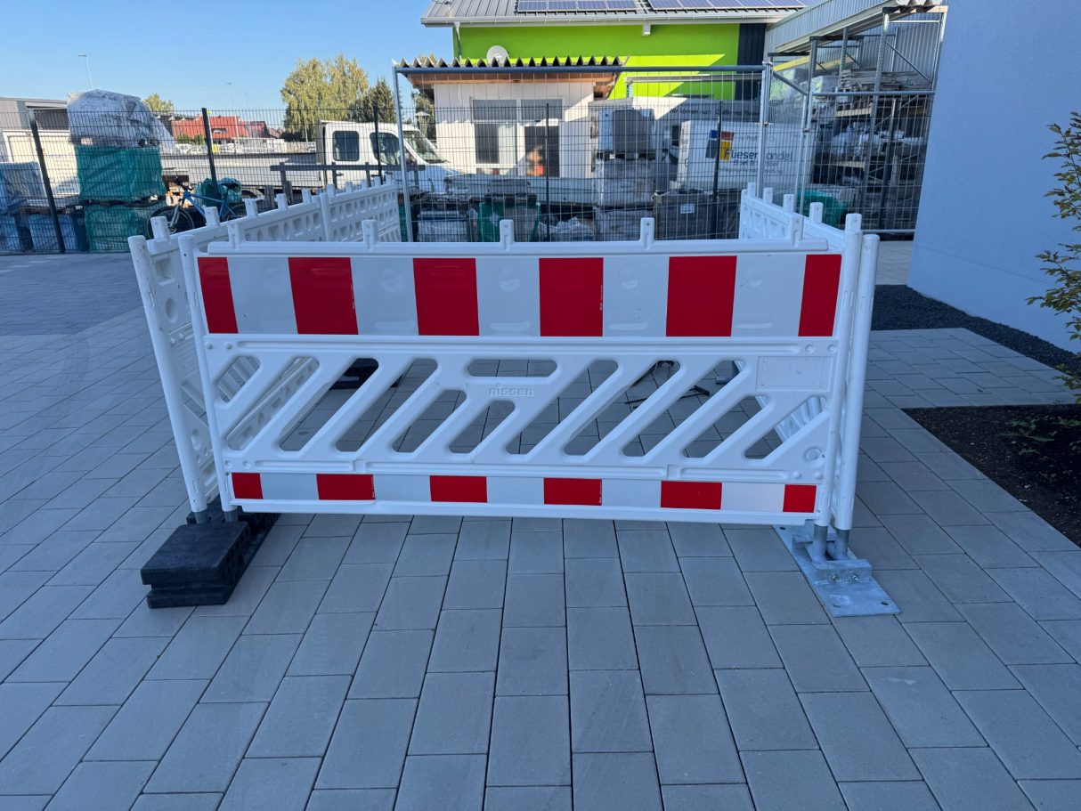 Weiße Absperrung mit roten Streifen, auf einem gepflasterten Boden.