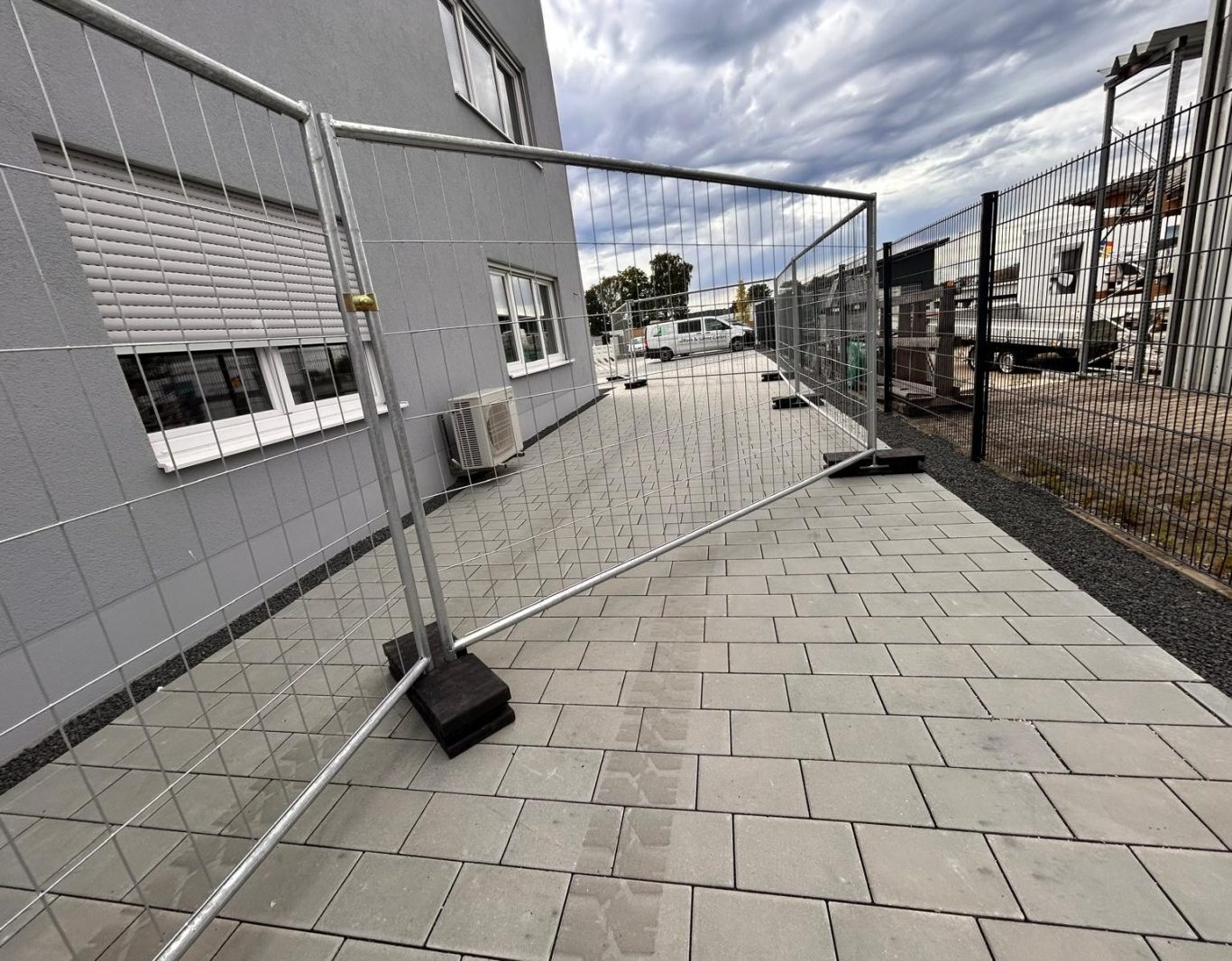 Bauzaun auf einem Gehweg neben einem Gebäude unter bewölktem Himmel. Mobile Defence - mobile Sicherheitszäune