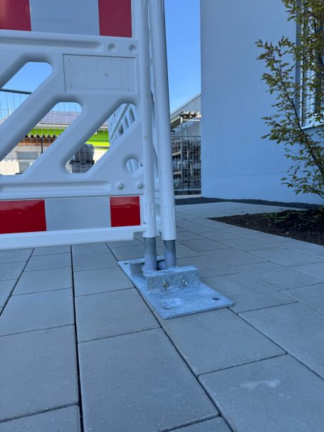 Bodenansicht einer Baustellenabsperrung mit Betonplatte und Pflanzen im Hintergrund.